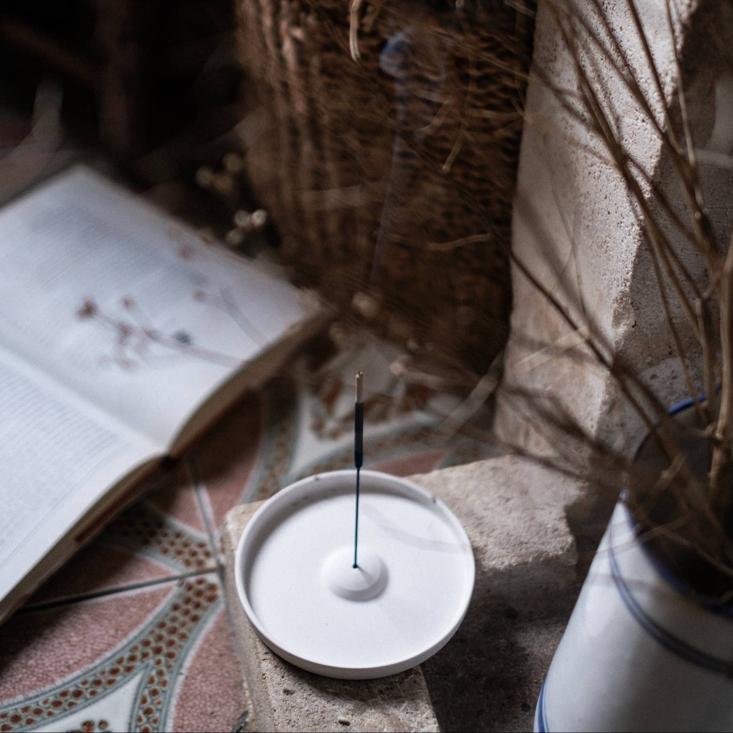 Incense stick holder with an incense stick on a textured surface with books and plants.