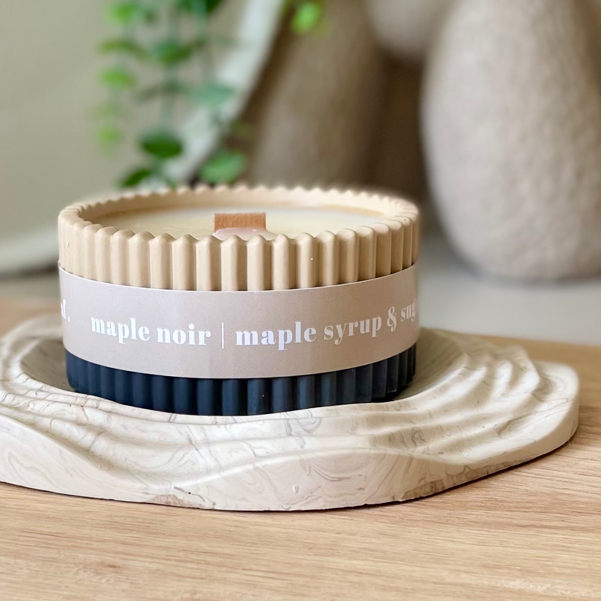 A ribbed jar candle with a beige top and bottom and a white paper band reading 'maple noir | maple syrup & soy', displayed on a stone coaster on a wooden surface with a softly blurred home background.