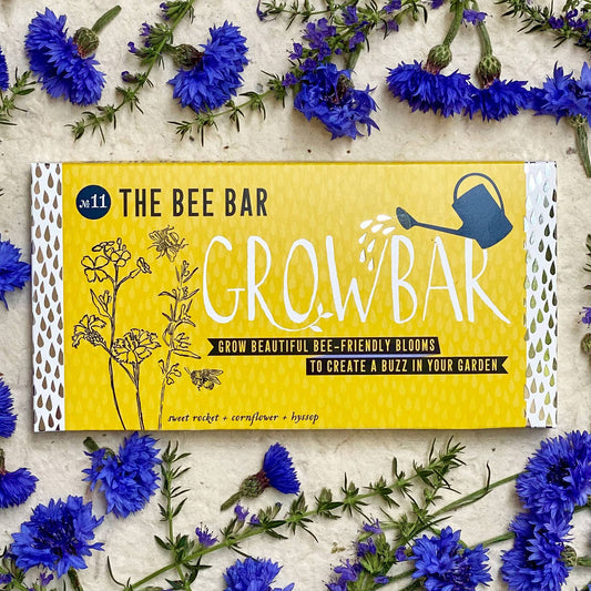A yellow rectangular package labeled 'The Bee Bar Grow Bar' lies on a light textured surface surrounded by scattered blue cornflowers. The package has white perforated side edges, black text, and simple line drawings of flowers, bees, and a watering can.
