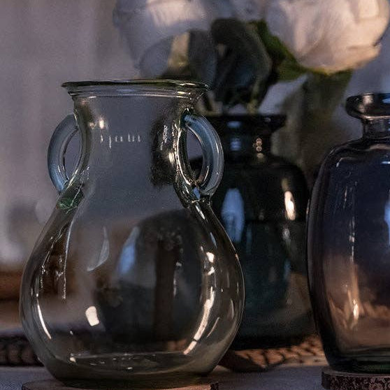 Three decorative glass vases with handles in smoky gray, dark blue-gray, and black, arranged on a table with woven coasters and white flowers in the background.