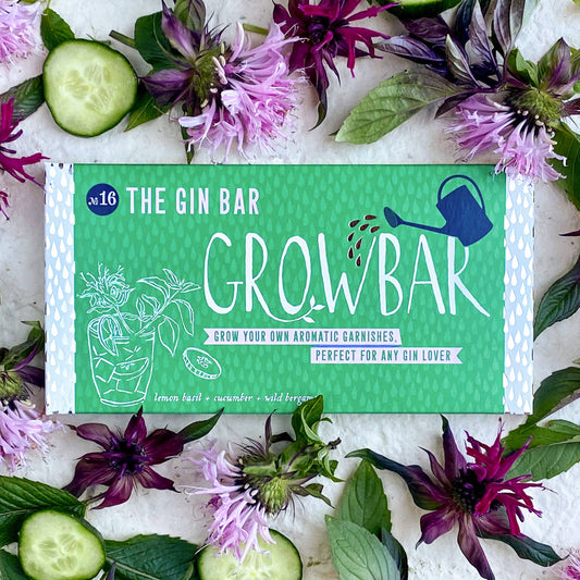 A rectangular green product box labeled 'THE GIN BAR GROWBAK' is laid flat on a white textured surface, surrounded by cucumber slices, purple flowers, and green leaves. The box has white text and simple line drawings, including a watering can graphic.