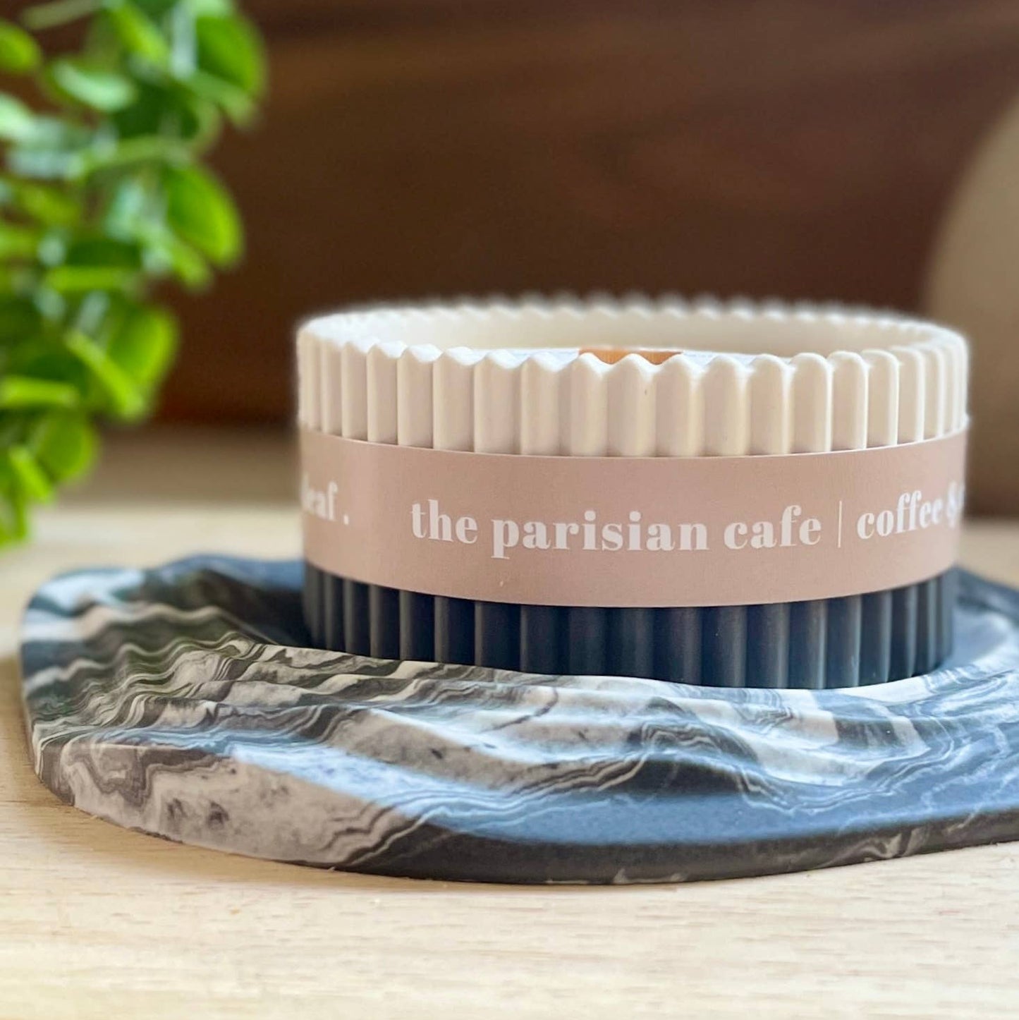 A round tin candle from The Parisian Cafe sits on a gray and white marbled coaster. The candle has a cream-colored fluted lid and a dusty pink paper band around the middle with white text.