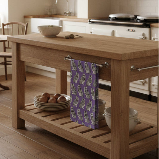 A tea towel with a repeating owl pattern on a lavender-purple background. The owls are white with black and gray striped details, arranged in a clean grid layout.